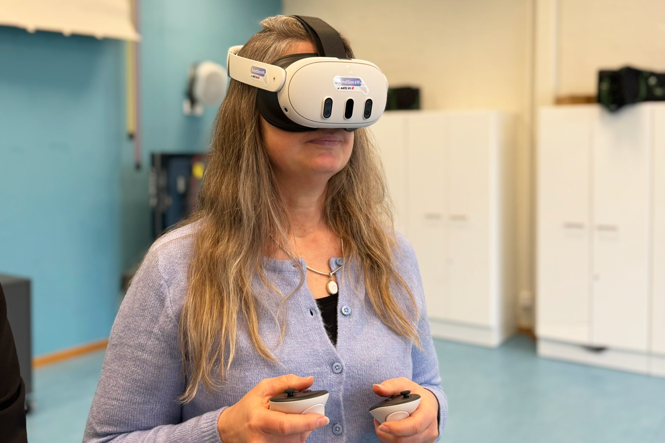 Kvinna i blå kofta och långt blondt hår som har VR-glasögon på sig. Bild.